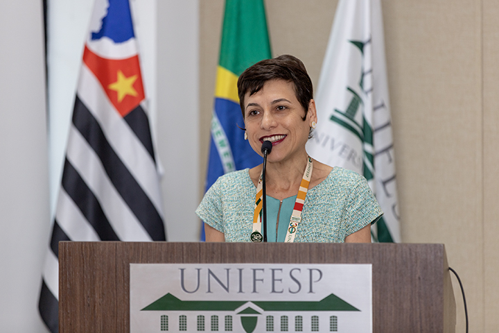 A imagem mostra uma mulher falando em um púlpito com o logo da UNIFESP (Universidade Federal de São Paulo) na frente. Ela está sorrindo e usando um crachá, o que indica que provavelmente está participando de um evento oficial ou conferência. Ela veste uma blusa clara de cor verde ou azul, e parece estar em um ambiente formal, possivelmente acadêmico.  Ao fundo, aparecem três bandeiras: a bandeira do estado de São Paulo, a bandeira do Brasil e uma bandeira institucional da UNIFESP.