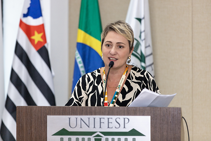 A imagem mostra uma mulher de cabelo curto e loiro falando em um púlpito, em um ambiente formal. Ela está usando um crachá colorido em volta do pescoço e um vestido com estampa preta e branca. O púlpito tem o logotipo da UNIFESP (Universidade Federal de São Paulo) na frente, e ao fundo estão visíveis as bandeiras do estado de São Paulo e do Brasil, além de outra bandeira institucional.