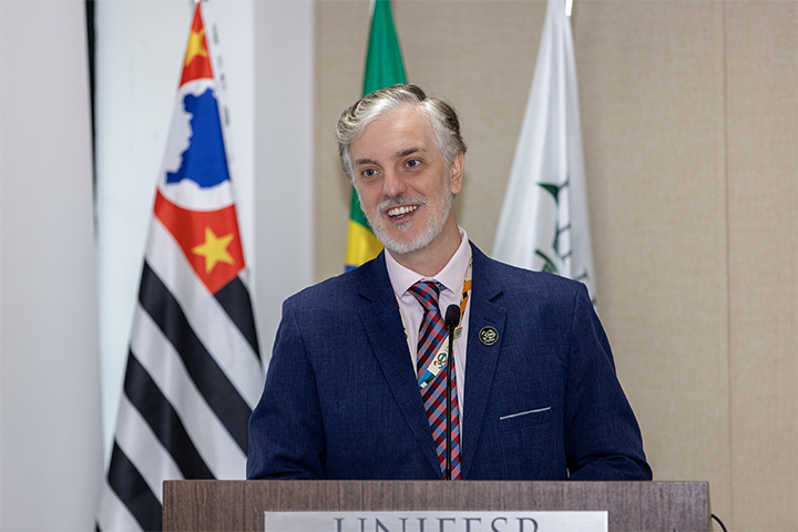 A imagem mostra um homem falando em um púlpito com o logo da UNIFESP (Universidade Federal de São Paulo) na frente. Ele está sorrindo, vestindo um terno azul escuro com gravata listrada, e também usa um crachá, sugerindo que está participando de um evento formal, possivelmente acadêmico ou científico. O homem parece estar no meio de um discurso ou apresentação, e o microfone está posicionado à sua frente.  Atrás dele, são visíveis três bandeiras: a bandeira do estado de São Paulo, a bandeira do Brasil e uma bandeira institucional da UNIFESP