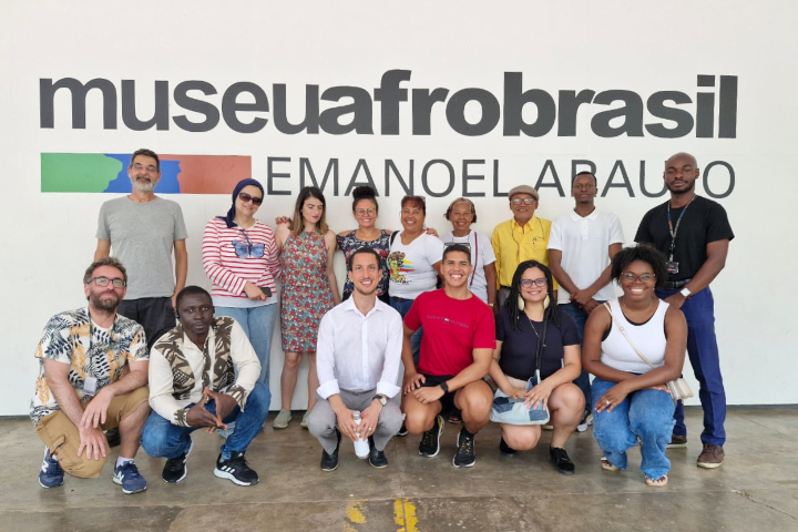 A foto mostra um grupo de 13 pessoas posando em frente à fachada do *Museu Afro Brasil Emanoel Araujo*, sorrindo e vestidas casualmente.