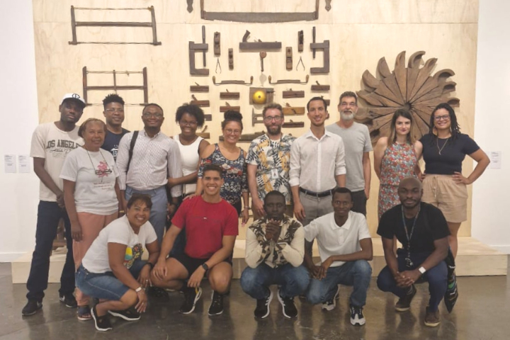 Um grupo de 15 pessoas diversas posa sorridente para uma foto em frente a uma parede com ferramentas antigas e elementos de madeira, no Museu Afro-Brasil Emanoel Araújo