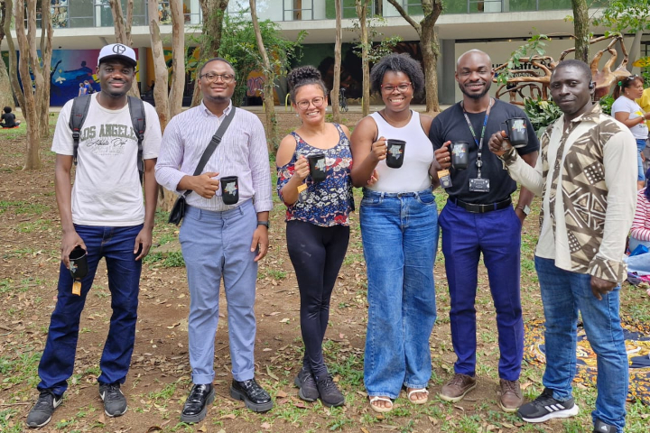 A foto mostra seis pessoas em um ambiente externo arborizado, sorrindo e segurando canecas pretas enquanto posam para a foto.