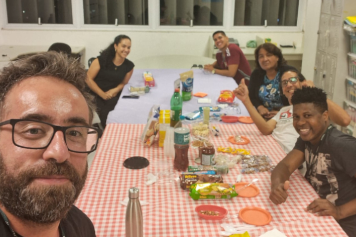 Sete pessoas sorriem em uma sala com uma mesa coberta por toalhas xadrez, cheia de alimentos, bebidas e itens de festa.