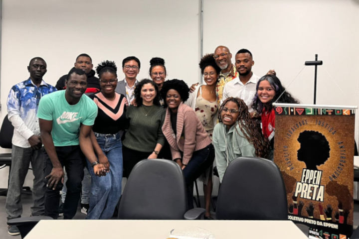 Um grupo de 12 pessoas diversas posa sorridente em uma sala, ao lado de um cartaz com a imagem de um perfil afro e o título "Eppen Preta".