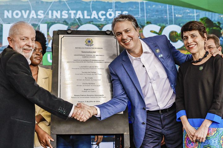 A imagem mostra uma cerimônia de inauguração com uma placa comemorativa. Três pessoas em destaque estão ao redor da placa, incluindo dois homens e uma mulher. Os dois homens estão apertando as mãos em frente à placa, ambos sorrindo, enquanto a mulher, ao lado direito deles, também sorri e apoia a mão no ombro do homem ao centro. Ao fundo, há um banner com as palavras parcialmente visíveis, indicando "Novas Instalações" e "Osasco", sugerindo que o evento está relacionado à cidade de Osasco, Brasil.