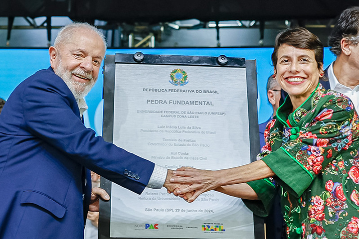 A imagem mostra outra cerimônia oficial de inauguração ou lançamento de pedra fundamental, onde duas pessoas em destaque, um homem e uma mulher, estão apertando as mãos em frente a uma placa comemorativa. O homem, à esquerda, está sorrindo e vestindo um terno azul escuro. A mulher, à direita, também sorri e está com uma roupa verde e colorida. A placa ao centro contém o título "Pedra Fundamental" e menciona o "Campus Zona Leste" da Universidade Federal de São Paulo (UNIFESP), além de listar autoridades presentes, incluindo o Presidente da República, o Governador de São Paulo, e o Ministro da Casa Civil.