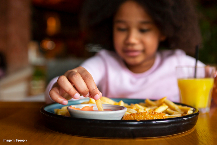 A imagem mostra uma criança sentada à mesa, pegando um pedaço de batata frita de um prato. O prato contém batatas fritas, nuggets e um molho rosado em um recipiente. Ao fundo, há um ambiente desfocado, indicando um possível restaurante ou lanchonete. À direita do prato, há um copo com suco de laranja. A criança está vestida com uma blusa clara e tem cabelo volumoso e encaracolado. A cena sugere um momento de refeição descontraído.   Legenda no canto inferior esquerdo: 