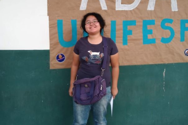 Na imagem, vemos uma pessoa sorridente posando em frente a um cartaz fixado em uma parede. Ela está usando uma camiseta roxa com uma ilustração de cachorro, calça jeans e uma bolsa roxa transversal. Também segura uma folha de papel na mão direita. Ao fundo, o cartaz tem letras grandes com parte da palavra "UNIFESP" visível, sugerindo um evento ou atividade relacionada à universidade. A pessoa parece estar feliz e confiante, possivelmente participando de algum tipo de evento acadêmico ou cultural.