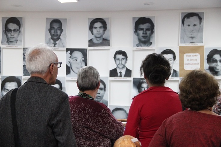 A imagem mostra um grupo de pessoas — quatro visíveis de costas — observando uma parede coberta por retratos em preto e branco de indivíduos, a maioria em estilo de foto de identidade. As pessoas na imagem parecem estar em uma exposição ou memorial, provavelmente relacionado a pessoas desaparecidas. À direita, há um cartaz com a pergunta: "ONDE ESTÃO OS NOSSOS DESAPARECIDOS?", reforçando a ideia de que se trata de uma homenagem ou ato de memória em relação a pessoas desaparecidas, possivelmente em um contexto de repressão política ou violência institucional. A faixa etária das pessoas presentes indica que podem ser familiares ou conhecidos das vítimas. O ambiente é fechado, bem iluminado e transmite um tom solene e reflexivo.