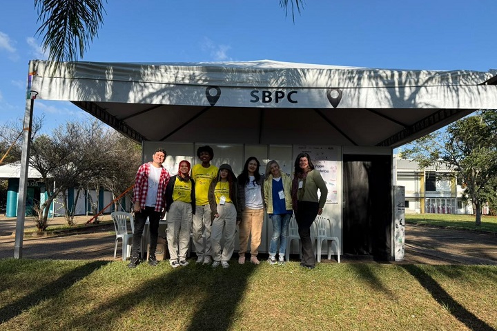 A imagem mostra um grupo de sete pessoas em frente a uma tenda branca com a inscrição SBPC (Sociedade Brasileira para o Progresso da Ciência), durante o dia. A tenda está montada em uma área externa, com gramado e árvores ao fundo, sob céu azul e tempo ensolarado.