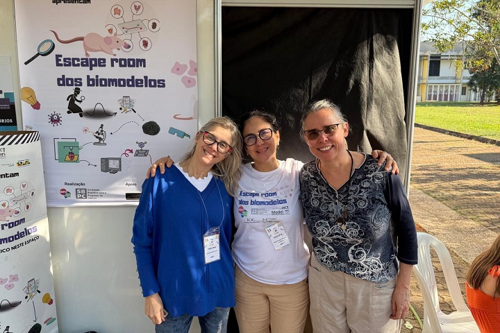 Três mulheres sorridentes posam em frente ao estande do projeto "Escape room dos biomodelos", atividade de divulgação científica realizada durante evento da SBPC, possivelmente no contexto da iniciativa Ciência Aberta do CNPEM.