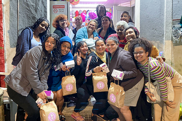 Grupo diverso de mulheres reunido na entrada de um local decorado, segurando kits de higiene e sorrindo para a câmera.