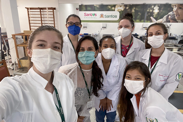 Seis mulheres de jaleco e máscara posam para uma selfie em um ambiente de saúde, representando profissionais da área.