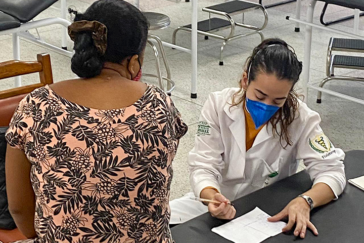 Uma profissional de jaleco branco e máscara azul escreve em um papel enquanto atende uma mulher de costas, vestindo uma blusa estampada. O ambiente parece ser um consultório ou espaço de atendimento.