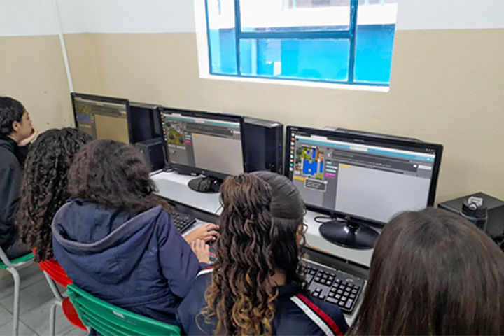 Um grupo de meninas está sentado em frente a computadores em uma sala de aula, programando em um ambiente gráfico de blocos coloridos. Elas estão concentradas, e a sala tem paredes claras e cadeiras coloridas.