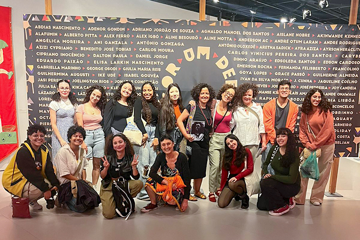 Um grupo diverso de mulheres posa sorrindo para a foto em frente a um mural com nomes de personalidades negras. O clima parece ser de confraternização e empoderamento.