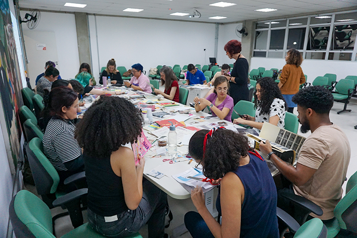 Um grupo de pessoas, a maioria mulheres, sentadas ao redor de uma grande mesa, envolvidas em uma atividade manual com recortes e materiais coloridos. O ambiente é uma sala de aula ou espaço de workshop.