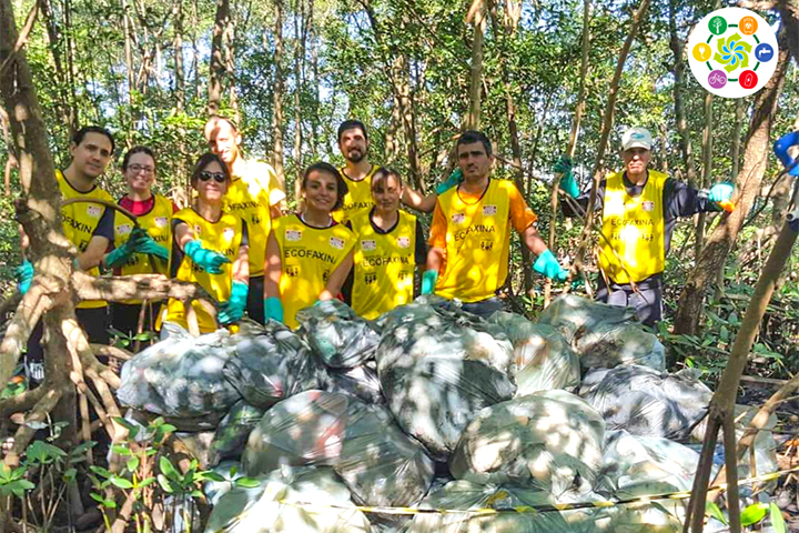 Voluntários com coletes amarelos do projeto EcoFaxina posam em área de mata ao lado de dezenas de sacos de lixo recolhido.