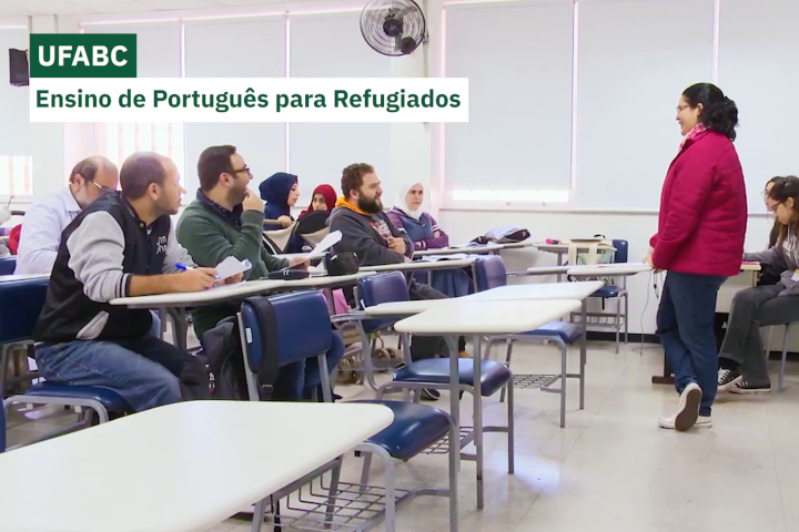 Sala de aula com adultos refugiados sentados em carteiras escolares, atentos à professora que fala à frente da sala. A professora veste casaco vermelho e todos parecem engajados na atividade.