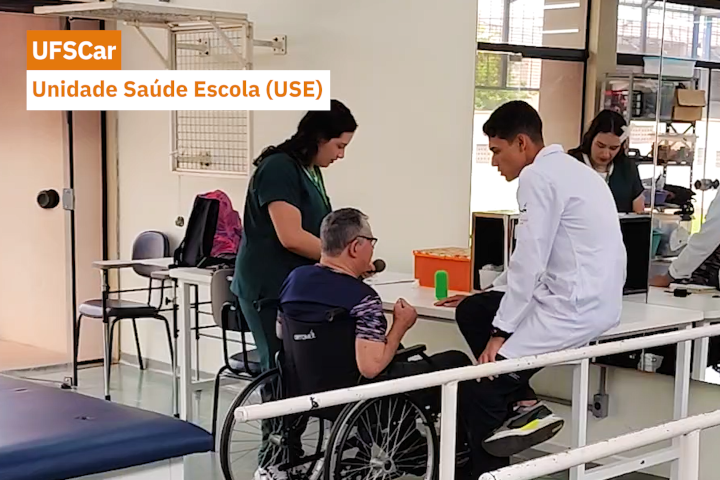 Espaço de atendimento em saúde com pessoas sendo acompanhadas por profissionais e estudantes. Um homem em cadeira de rodas conversa com um jovem de jaleco branco, enquanto outras pessoas trabalham ao fundo.