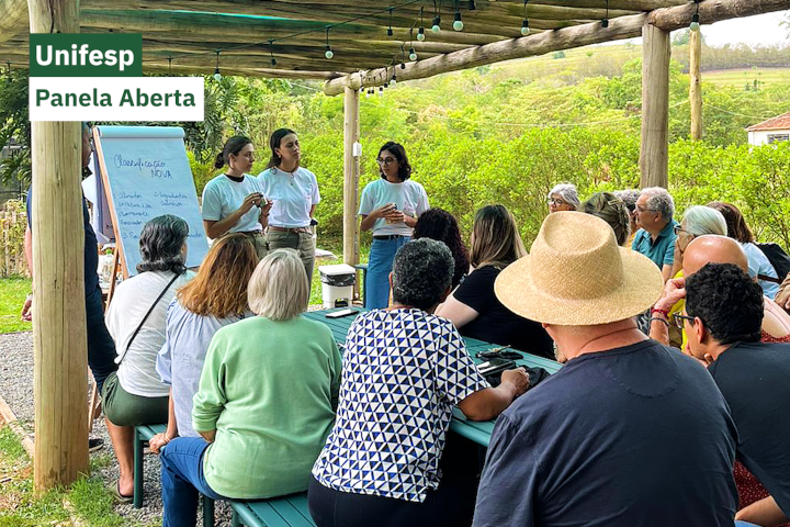 Encontro ao ar livre sob um pergolado, com pessoas sentadas ao redor de uma mesa de madeira. Três jovens apresentam um conteúdo escrito em um quadro branco, em meio a um cenário natural com árvores ao fundo.