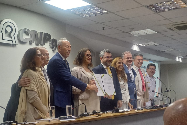Um grupo de pessoas, algumas sorrindo, posa atrás de uma mesa em um evento formal, com documentos sendo assinados.