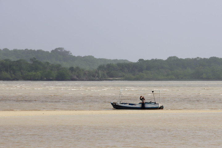 Um barco pequeno com uma pessoa a bordo está em águas rasas, com árvores ao fundo sob um céu nublado.