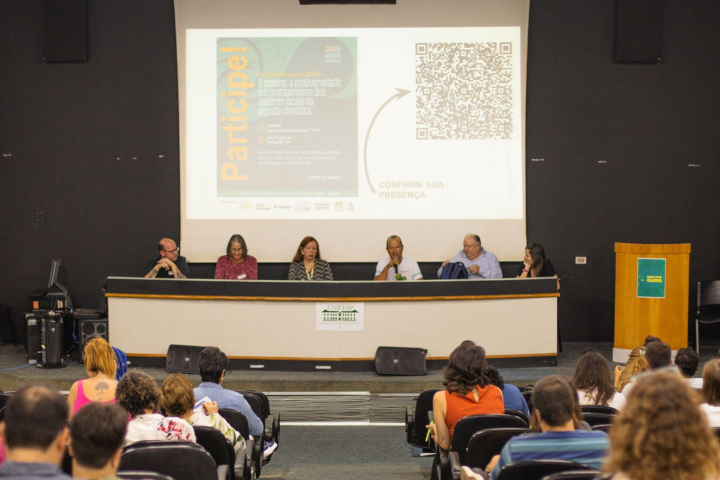 Uma mesa com palestrantes de frente para uma plateia em um auditório, com uma projeção de slides no telão.