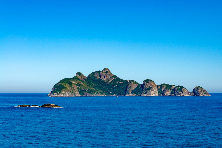 Uma ilha rochosa e verdejante se destaca em um vasto oceano azul sob um céu claro.
