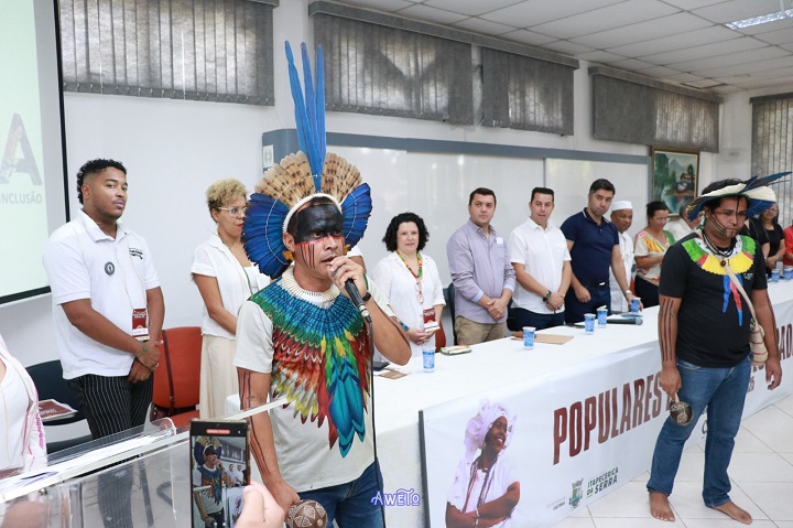A imagem mostra um evento formal com foco na valorização de saberes populares, indígenas e afrodescendentes. Em destaque, dois homens indígenas com cocares e pintura corporal participam da cerimônia, um deles falando ao microfone. Ao fundo, uma mesa com autoridades e organizadores em pé, diante de uma faixa com a palavra “POPULARES”. O ambiente é institucional, sugerindo um encontro cultural promovido por um órgão público.