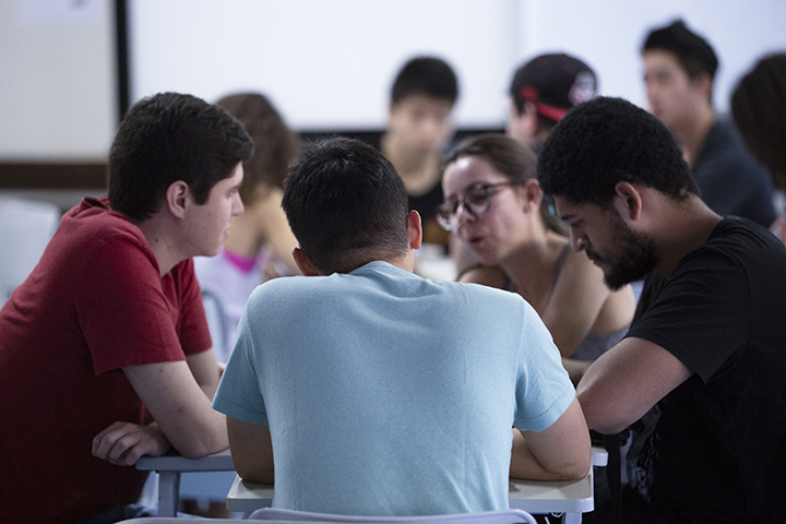 A imagem mostra um grupo de jovens reunidos em uma sala de aula ou ambiente acadêmico, envolvidos em uma discussão ou atividade colaborativa. No primeiro plano, três pessoas estão sentadas ao redor de uma mesa, inclinadas umas para as outras, sugerindo uma conversa envolvente. Uma mulher de óculos está falando, enquanto os outros parecem ouvir atentamente.  Ao fundo, outros estudantes podem ser vistos, alguns desfocados, reforçando a ideia de um ambiente de aprendizado ou trabalho em grupo. A atmosfera sugere cooperação e troca de ideias, possivelmente em um contexto educacional ou universitário.