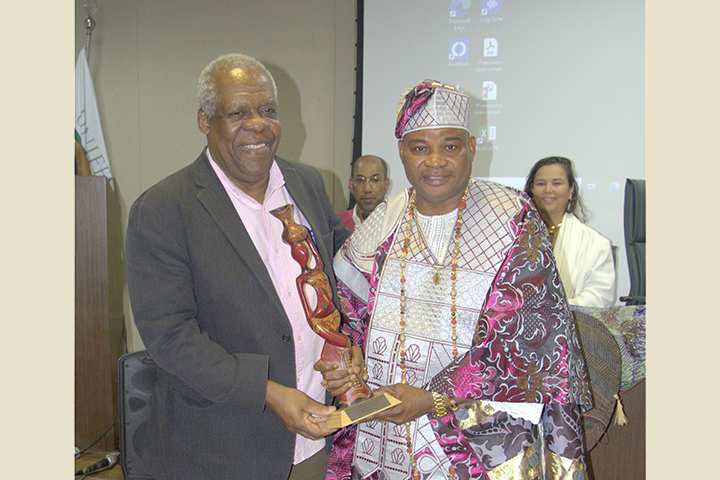 A imagem mostra um momento de premiação durante um evento formal. Um homem negro de cabelos grisalhos, vestindo um blazer escuro e camisa rosa, entrega uma escultura ao Oba Adekunlé Aderonmu, que está trajado com vestes tradicionais iorubás luxuosas. Ambos sorriem para a câmera. Ao fundo, há uma mulher (possivelmente organizadora ou autoridade) também sorrindo, sentada à mesa principal, em frente a uma tela de projeção com ícones de computador visíveis. A cena transmite um clima de celebração e reconhecimento cultural.