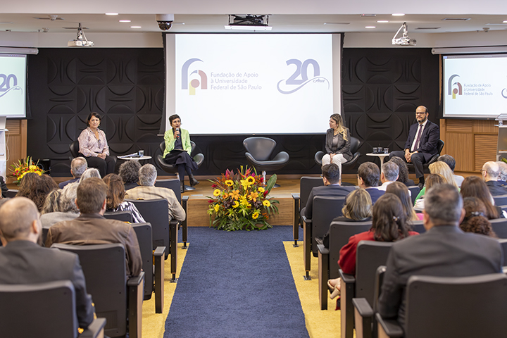 A imagem mostra um evento formal realizado em um auditório. No palco, há cinco pessoas sentadas em cadeiras, participando de uma mesa-redonda ou painel de discussão. O ambiente é bem iluminado e o público está atento, com muitas pessoas sentadas em poltronas acolchoadas, voltadas para o palco. No fundo do palco, há duas telas grandes projetando os logotipos da Fundação de Apoio à Universidade Federal de São Paulo (FapUnifesp) e a marca de 20 anos da fundação. Entre os palestrantes, há uma decoração floral central com flores amarelas, vermelhas e verdes. O evento parece estar relacionado a uma celebração institucional ou conferência da FapUnifesp, possivelmente comemorando seus 20 anos de atuação. A atmosfera é profissional e solene, com os participantes vestidos formalmente.