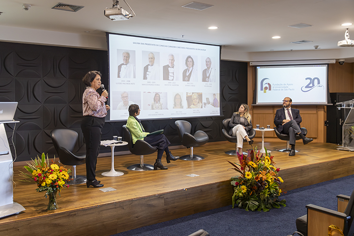 Nesta imagem, vemos a continuação do mesmo evento formal mostrado anteriormente, realizado em um auditório. A cena foca no palco, onde uma das participantes (à esquerda) está de pé, segurando um microfone e falando ao público. Ela está ao lado de um púlpito com um arranjo de flores coloridas na base.  No fundo do palco, há uma projeção com a galeria dos presidentes do Conselho Curador da Fundação de Apoio à Universidade Federal de São Paulo (FapUnifesp), com fotos e nomes de diversos ex-presidentes e membros honorários.  Sentadas no palco, outras três pessoas acompanham a apresentação: uma mulher de verde, uma mulher de bege e um homem de terno escuro. Eles estão em cadeiras modernas, voltadas para a apresentadora. A identidade visual da FapUnifesp e o logotipo de 20 anos continuam visíveis nas telas