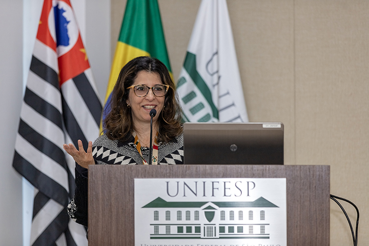 Mulher de óculos, cabelos soltos, sorri enquanto fala ao microfone no púlpito da Unifesp, com bandeiras ao fundo.