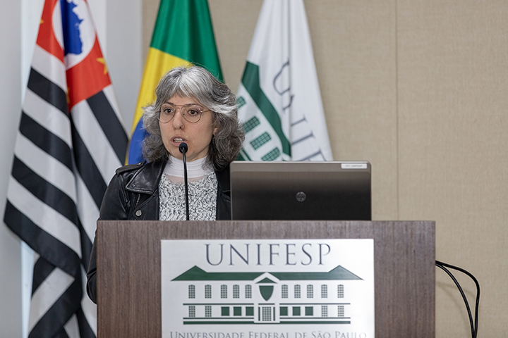 Mulher de cabelos grisalhos e óculos fala ao microfone, atrás de púlpito com logotipo da Unifesp e bandeiras.