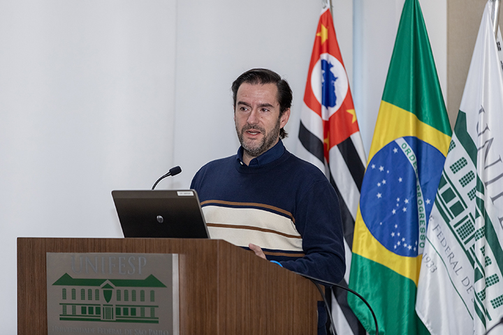 Homem de barba e cabelos escuros fala ao microfone, com bandeiras do Brasil, São Paulo e Unifesp atrás.
