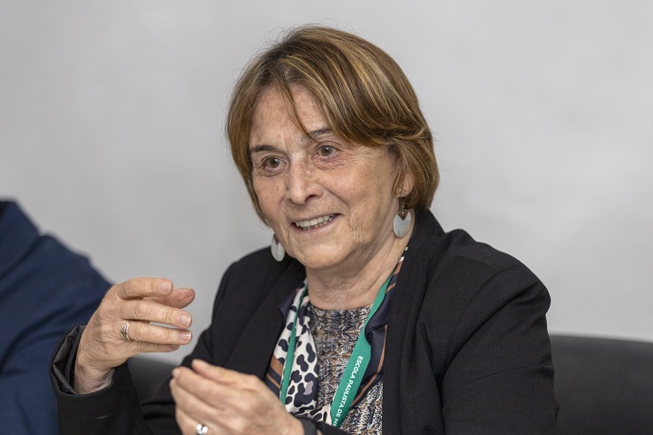 Na imagem, vemos uma mulher mais velha com cabelos curtos e castanhos. Ela está sorrindo e gesticulando com as mãos, parecendo estar envolvida em uma conversa ou explicação. Veste um blazer escuro sobre uma blusa estampada e usa brincos arredondados. Também está usando um crachá verde no pescoço, sugerindo que está em um evento, conferência ou ambiente profissional. O fundo é neutro e desfocado, mantendo o foco na pessoa retratada.