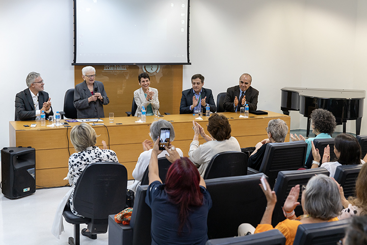 A imagem mostra um evento em uma sala de conferências, onde cinco pessoas estão sentadas atrás de uma mesa, participando de um painel. Eles parecem estar sendo aplaudidos pelo público, que está sentado em cadeiras organizadas em fileiras. Algumas pessoas na plateia estão tirando fotos com seus celulares. No fundo, há uma tela de projeção e um piano de cauda.
