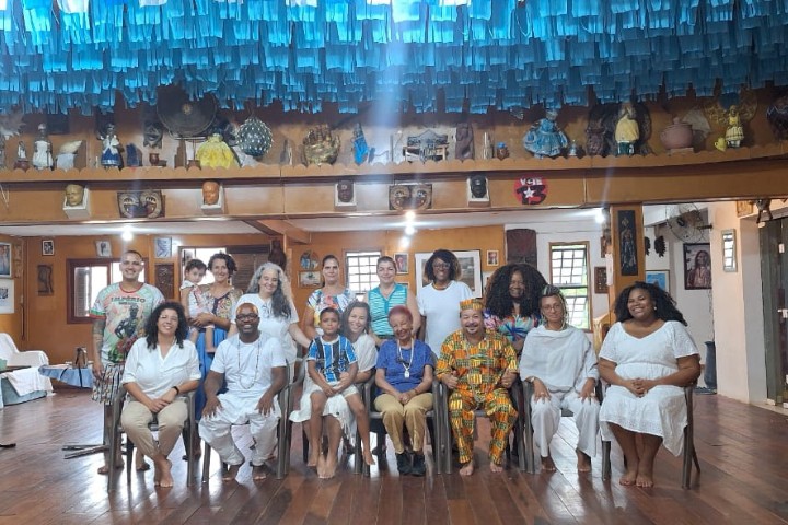 A imagem mostra um grupo diverso de pessoas reunidas em um espaço cultural decorado com elementos afro-brasileiros. Muitas estão vestidas de branco, simbolizando tradições de matriz africana. O ambiente e os trajes sugerem uma celebração ou encontro de caráter cultural ou religioso.