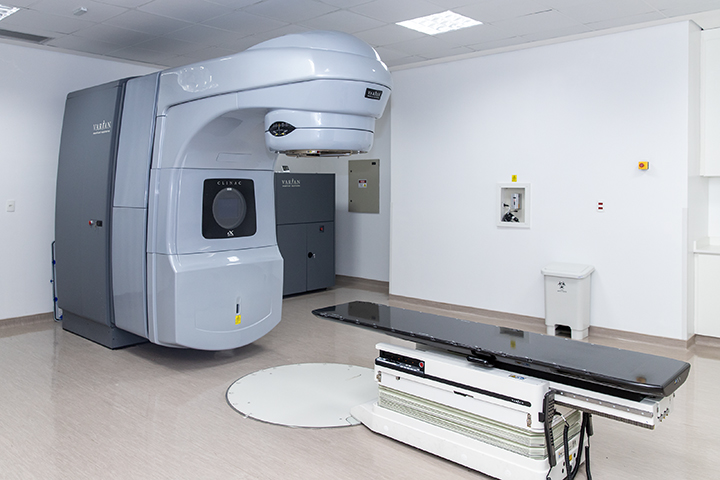 A imagem mostra uma sala clínica equipada com um acelerador linear (marca Varian), utilizado em tratamentos de radioterapia para pacientes com câncer. O equipamento é de grande porte, fixado ao chão e ao teto, com uma mesa móvel posicionada em frente a ele, onde o paciente é colocado durante o procedimento.  O ambiente é limpo, bem iluminado e estéril, como é padrão em instalações médicas desse tipo. A sala também conta com itens de segurança e controle, como painéis na parede, uma lixeira hospitalar e revestimento próprio para áreas de radiologia.