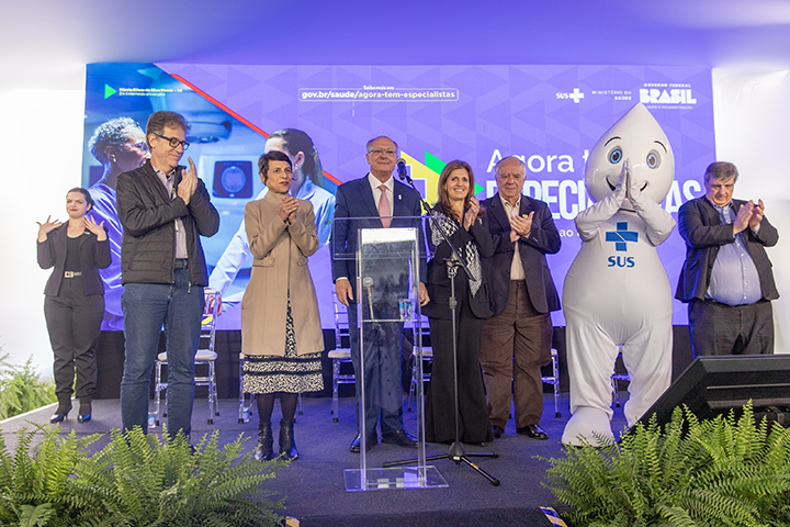 A imagem mostra um evento oficial com sete pessoas no palco, incluindo um mascote do SUS (Sistema Único de Saúde) vestido de branco com uma cruz azul. Ao centro, há um púlpito com microfones. As pessoas estão aplaudindo e sorrindo. O fundo exibe painéis institucionais com logotipos do governo federal do Brasil e do Ministério da Saúde, destacando o SUS. Há também plantas decorativas na frente do palco.