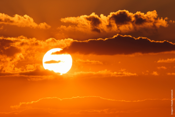 A imagem mostra um pôr do sol deslumbrante com tons quentes de laranja e dourado. O sol aparece parcialmente coberto por nuvens escuras, criando um efeito dramático e contrastante. As nuvens superiores estão iluminadas nas bordas, destacando-se contra o céu intenso e vibrante. A composição transmite uma sensação de calor, tranquilidade e beleza natural.