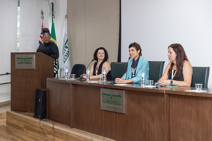 A imagem retrata um painel de discussão em um evento na Universidade Federal de São Paulo (UNIFESP). Três mulheres estão sentadas atrás de uma mesa com microfones e garrafas de água, enquanto uma quarta mulher, vestida de preto, está de pé atrás de um púlpito, aparentemente conduzindo a sessão. A mulher no centro da mesa, vestindo um blazer azul, sorri enquanto fala, enquanto as outras participantes ouvem atentamente. Ao fundo, bandeiras, incluindo a do Brasil e a do estado de São Paulo, reforçam o caráter institucional do evento. O ambiente sugere um debate acadêmico ou científico.