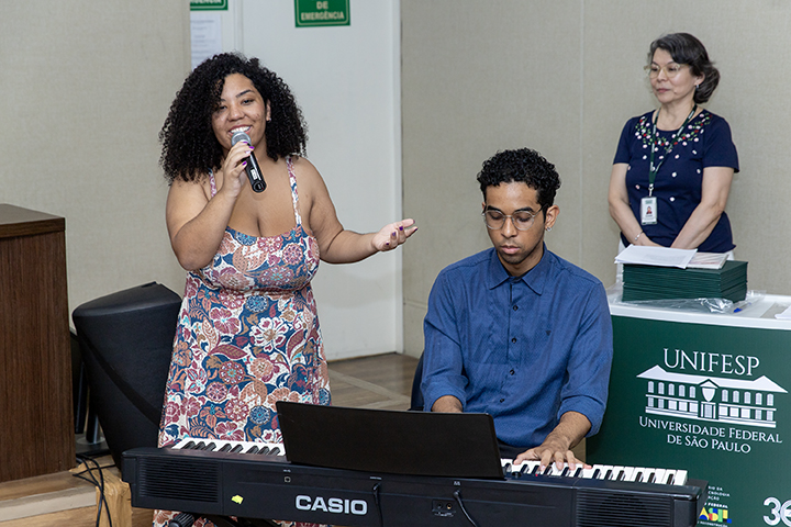 A imagem mostra uma apresentação musical em um evento na Universidade Federal de São Paulo (UNIFESP). Uma mulher com cabelos cacheados, vestindo um vestido estampado, canta ao microfone com uma expressão animada. Ao lado dela, um homem usando óculos e uma camisa azul toca um teclado da marca Casio, concentrado na música. Ao fundo, uma mulher com crachá observa a apresentação perto de uma mesa com materiais da UNIFESP, indicando um ambiente institucional. A cena sugere um momento cultural dentro de um evento acadêmico.