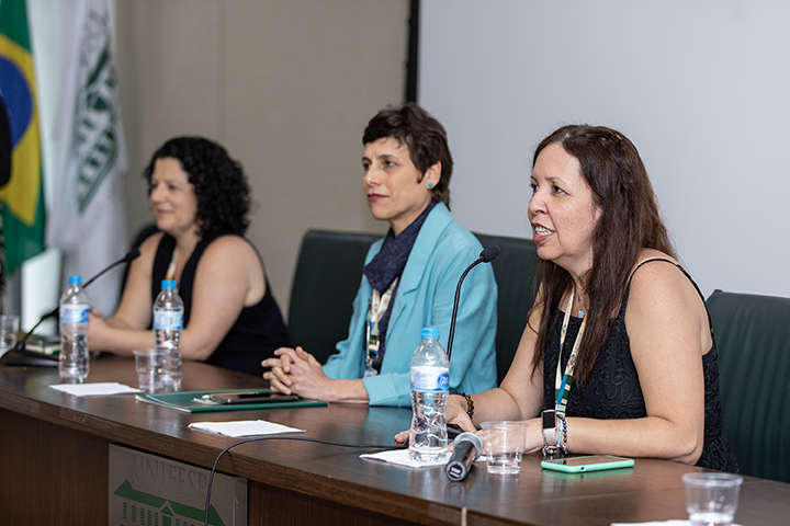 A imagem mostra três mulheres sentadas em uma mesa durante um evento acadêmico na Universidade Federal de São Paulo (UNIFESP). A mulher à direita, de cabelos longos e vestindo preto, está falando ao microfone com uma expressão engajada. Ao seu lado, uma mulher de blazer azul escuta atentamente, sorrindo levemente, enquanto a terceira participante, vestindo preto e com cabelos cacheados, observa a discussão. Na mesa, há garrafas de água, celulares e materiais de anotação. Bandeiras do Brasil e da UNIFESP aparecem ao fundo, reforçando o caráter institucional do evento.