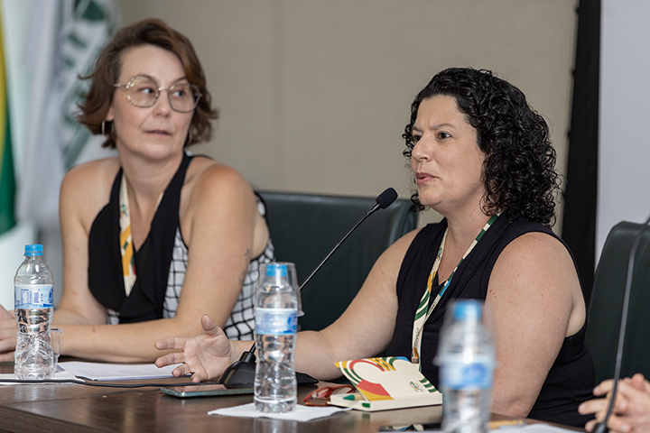 A imagem mostra duas mulheres sentadas em uma mesa durante um evento ou conferência. A mulher à direita está falando ao microfone, gesticulando com a mão, e parece engajada na conversa. Ela tem cabelos cacheados escuros e veste uma blusa preta sem mangas. A mulher à esquerda, que usa óculos e tem cabelos curtos e ondulados, observa atentamente, com uma expressão séria. Ambas usam crachás e estão acompanhadas de garrafas de água, cadernos e canetas sobre a mesa. O ambiente sugere um evento formal, possivelmente um painel ou discussão acadêmica, com bandeiras ao fundo.
