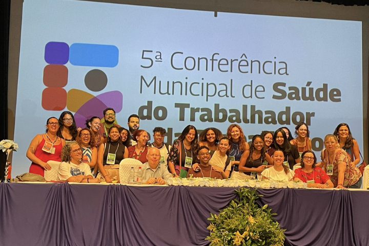 A imagem mostra um grupo diverso de pessoas reunidas em um palco durante a "5ª Conferência Municipal de Saúde do Trabalhador e da Trabalhadora". Ao fundo, há um telão com o nome do evento e um logotipo colorido. As pessoas estão sorrindo, sugerindo um momento de confraternização ou encerramento da conferência.