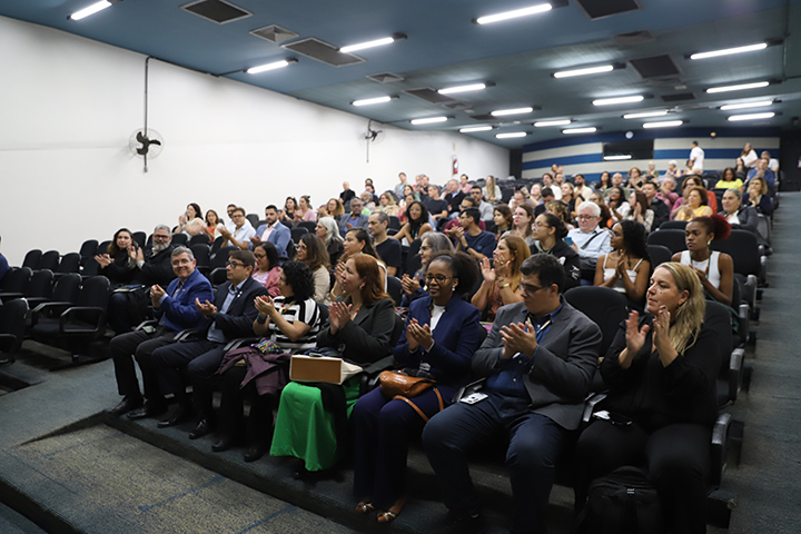 A imagem mostra a plateia de um auditório cheia, composta por diversas pessoas sentadas e aplaudindo. O ambiente é bem iluminado com luzes no teto, e as pessoas estão vestidas formalmente, sugerindo um evento acadêmico ou institucional. A disposição das cadeiras e a concentração das pessoas indicam um momento de celebração ou solenidade, possivelmente relacionado à imagem anterior de uma cerimônia na Universidade Federal de São Paulo (UNIFESP).