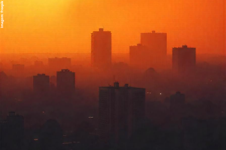 A imagem mostra uma cidade envolta em uma névoa densa, possivelmente causada pela poluição do ar. O céu tem tons alaranjados e avermelhados, sugerindo que a foto foi tirada durante o nascer ou o pôr do sol. Os edifícios altos aparecem como silhuetas escuras contra o fundo luminoso, criando um efeito dramático e contrastante. A atmosfera carregada e a visibilidade reduzida indicam a presença de partículas no ar, o que pode ser um indicativo de problemas ambientais relacionados à qualidade do ar na cidade.